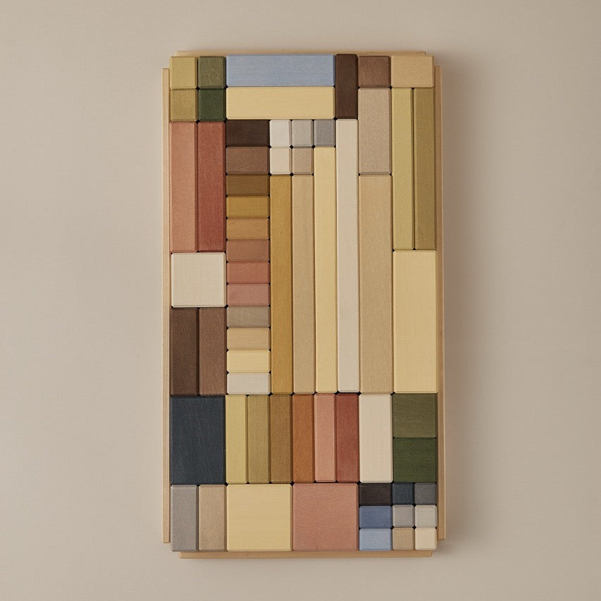 A rectangular wall-mounted art piece composed of many stacked painted blocks in earthy tones of beige, brown, cream, muted blue, green, and terracotta, shown against a plain warm beige wall.