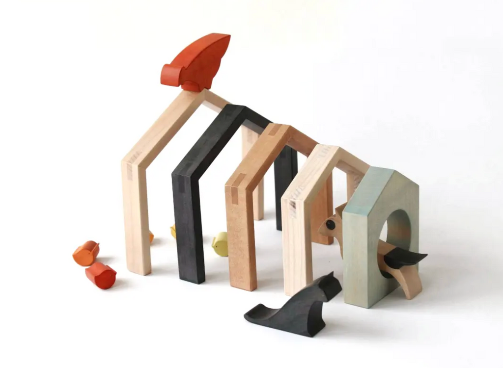A set of wooden architectural toy blocks and small sculptural pieces arranged on a white studio background. The pieces include natural wood arches, black and reddish-brown blocks, and a pale mint green arch, with a black cat-shaped piece placed in front.