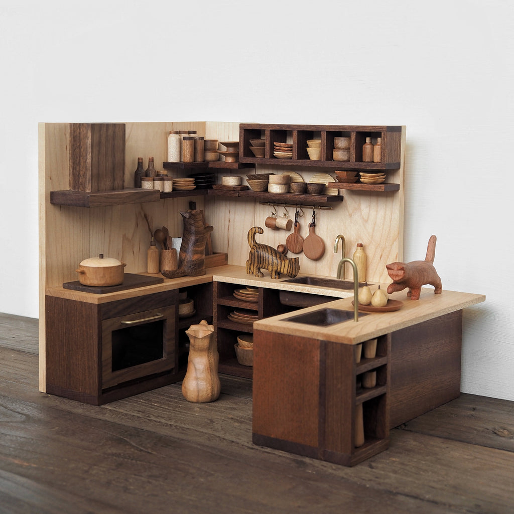 A miniature wooden dollhouse kitchen set with dark brown cabinets, light natural wood countertops and backsplash, and small toy kitchen accessories including pots, bowls, a sink, and a cat figurine, displayed on a wooden floor against a white wall.