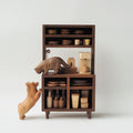A small wooden display cabinet with shelves and cubbies, styled with miniature tabletop accessories including cups, plates, and carved animal figures, photographed against a clean light gray studio background.